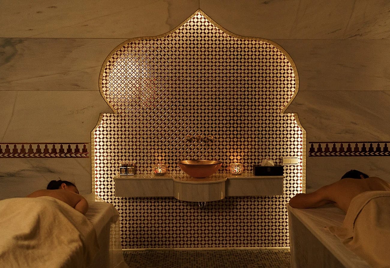 Two guests resting on marble beds in front of a glowing tiled arch in a private hammam