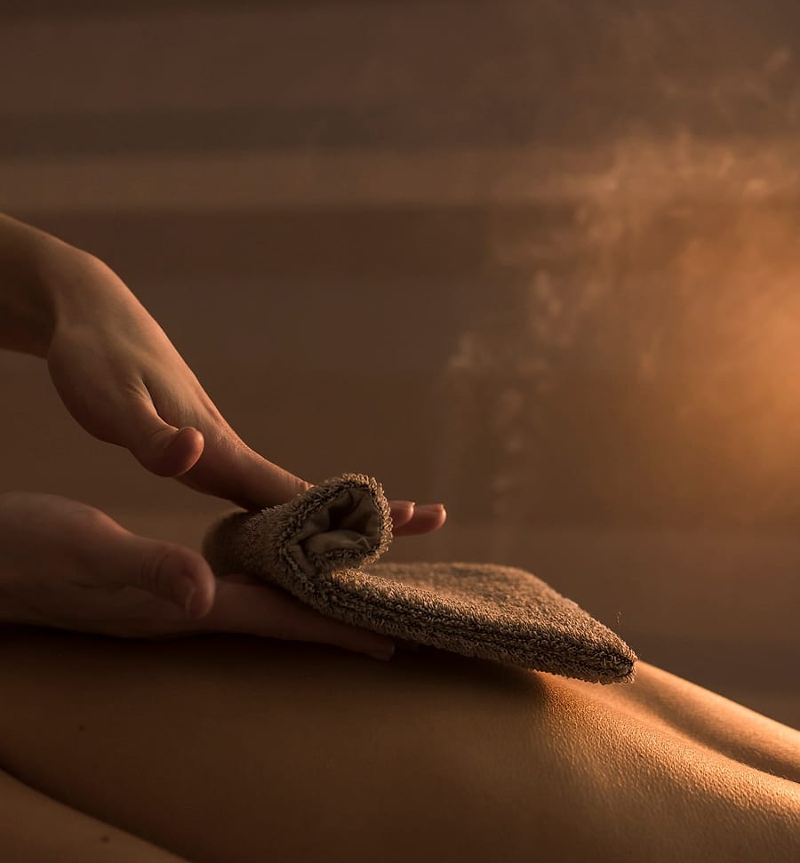 A therapist gently wipes a warm towel mitt over a guest’s back in the steam